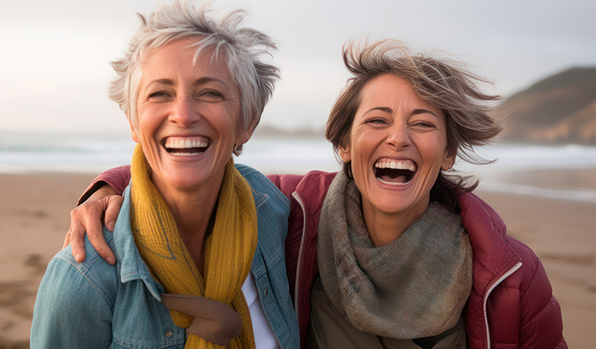 Zwei Frauen halten sich am Strand im Arm und lachen in die Kamera.