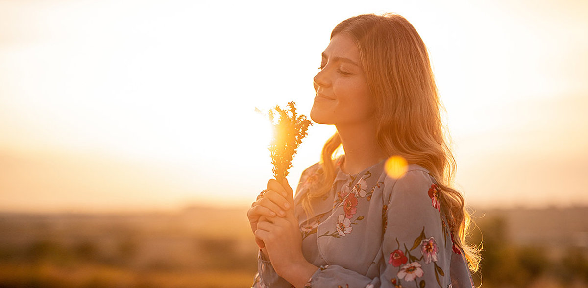 Junge Frau mit langen welligen Haaren in der Abendsonne hält Kräuterzweige in der Hand und lächelt